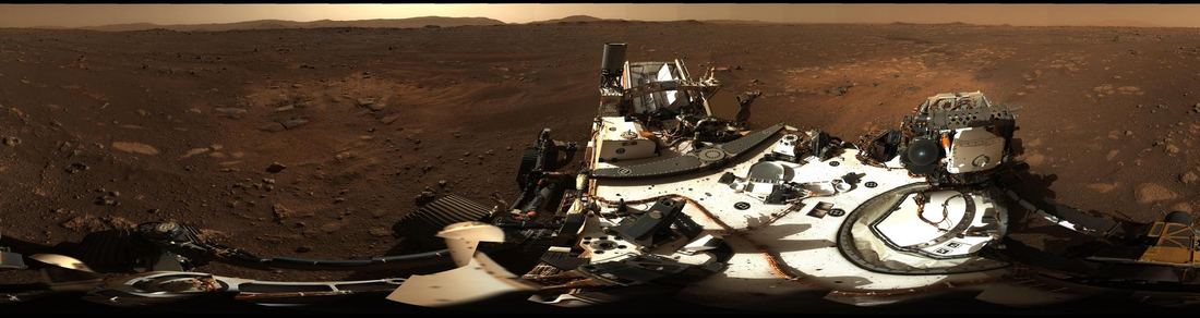 A wide panorama of rocky, dusty ground inside Jezero Crater on Mars under a hazy sky